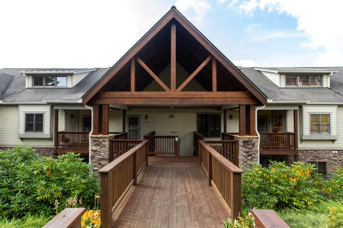 A welcoming wooden entrance ramp leads to a home framed by stone columns and lush greenery. The view hints at the promise of cozy gatherings and leisurely afternoons on the porch, surrounded by scenic outdoors.