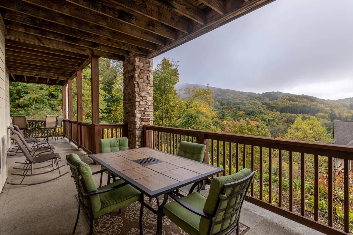 The image showcases a spacious exterior deck at Caribou Crossing, featuring a stylish dining table with green chairs set against a backdrop of sturdy wooden railings and stone pillars. Beyond, breathtaking mountain views invite guests to relax, dine al fresco, and soak in the natural beauty of Banner Elk.