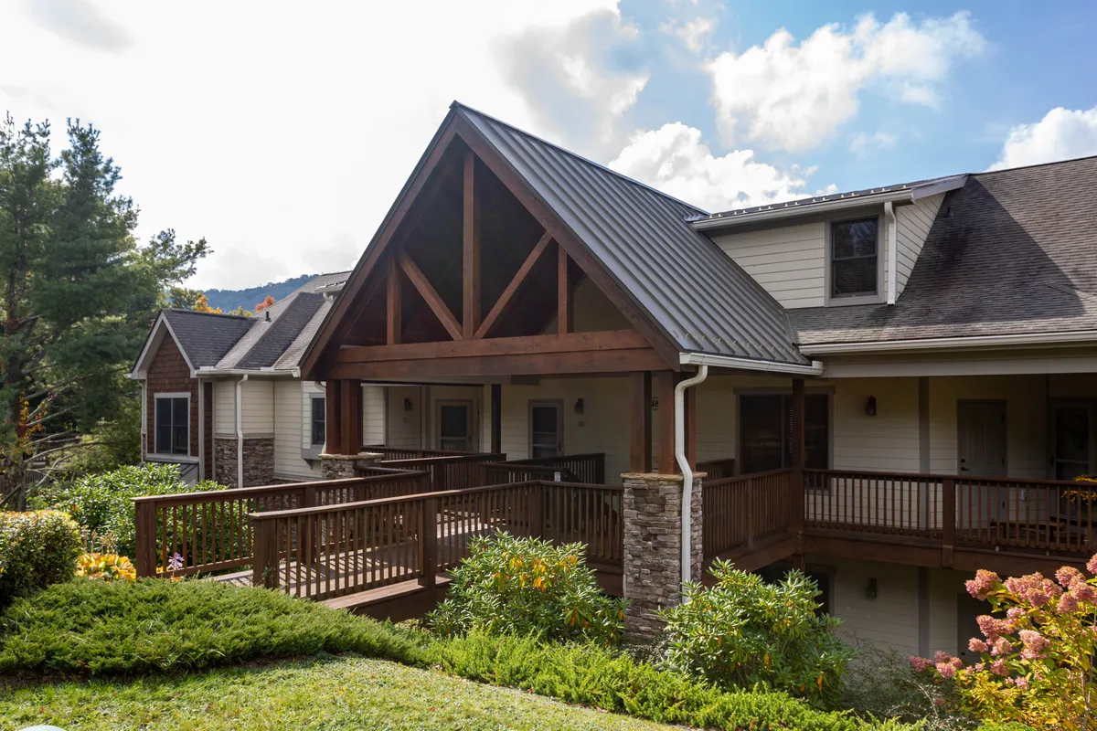 This image displays a broad wooden wrap-around porch accented with stone pillars and a dramatic triangular roof structure. The porch is surrounded by lush landscaping, suggesting a serene connection to nature. The expansive setup clearly highlights the generous outdoor seating area, perfect for gatherings or quiet moments alike.
