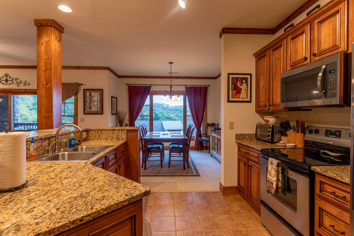 A bright and cozy kitchen-dining area, featuring granite countertops, wooden cabinets, a dining table set before a large window with scenic views. The space is designed for cooking together and enjoying shared meals.