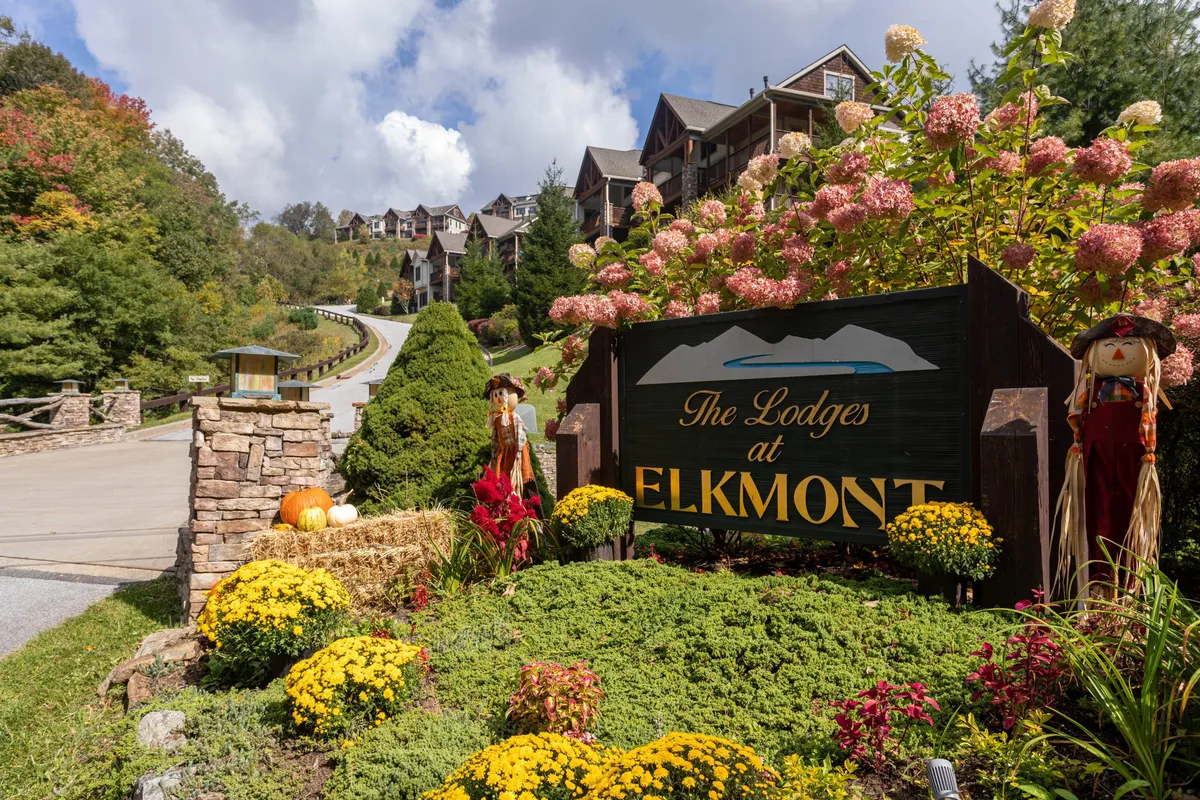 The image showcases the welcoming entrance signage for 'The Lodges at Elkmont,' surrounded by colorful flowerbeds, playful scarecrows, and detailed stonework. The bright and well-maintained exterior conveys the inviting atmosphere of the resort community, set against a mountain backdrop.