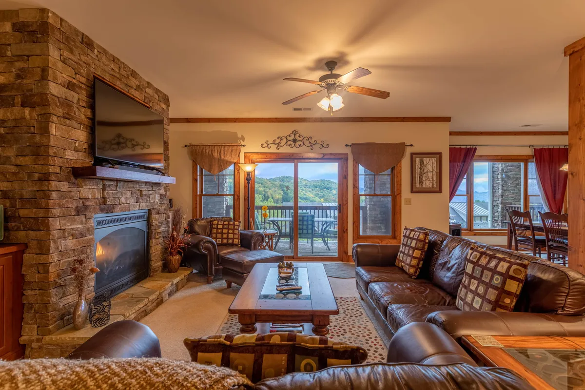 The image displays a spacious open living area with a prominent stacked stone fireplace, large windows offering scenic outdoor views, plush leather sofas, and a substantial wooden dining table. The light-filled space exudes warmth and invites gatherings around the fire or the table.