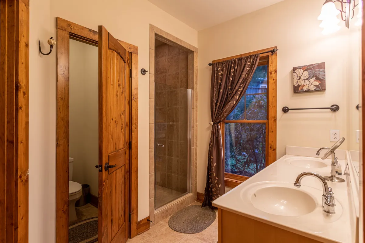 A serene, modern bathroom with double vanity sinks, a spacious shower area, and a soft curtain-filtered window. The space is designed for relaxation and rejuvenation.