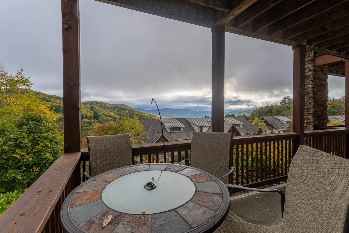 A patio table set is positioned to overlook sweeping mountain views, framed by vibrant foliage. The setup is intimate and ideal for either a morning coffee or an evening toast. The overall mood is tranquil, emphasizing the connection to nature and the picturesque landscape.